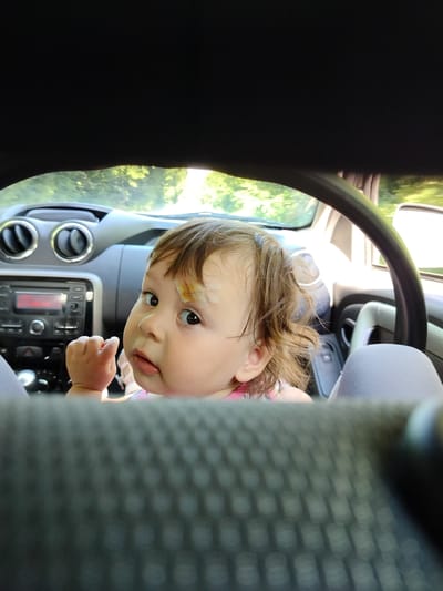 In this close-up, medium shot, a young child sits in the passenger seat of a car, looking directly at the camera. The child has brown hair, and a small bandage is visible on their forehead. Their expression is curious and slightly wary, with eyes wide open and mouth slightly ajar. The child's left hand is raised and clenched.
The car's interior is visible, with the dashboard and steering wheel in the background. The dashboard features a car radio and air vents. The scene appears to be set during the daytime, with sunlight filtering through the car windows, illuminating the child's face and hair. The background outside the car suggests a natural environment with greenery, possibly trees and foliage.
The image's composition is framed by the car's interior, with dark elements in the foreground and top obscuring the full view. The focus is sharply on the child, with the car's interior elements slightly blurred. The overall impression is one of an intimate and candid moment, capturing the child's presence within the car. The provided location context is Степное Полеологово, Russia. No visible text is present in the image.