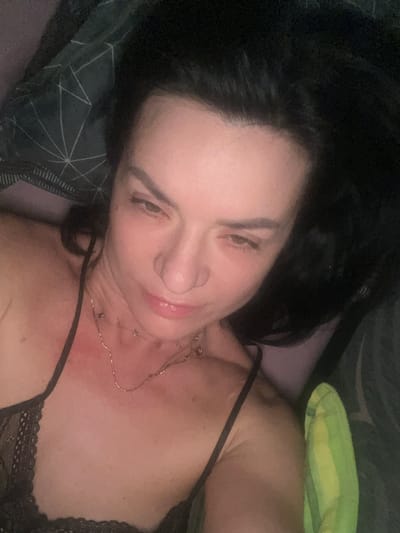 A medium shot captures a woman indoors, lying down. She has dark hair and is wearing a black lace top with spaghetti straps and a gold necklace. She is lying on a patterned dark green and gray pillow. Another pillow is to her right. The scene location is the Smederevska Palanka Municipality, Serbia.