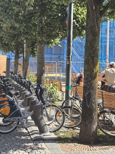 The image depicts a row of rental bicycles parked along a tree-lined street in Lund, Sweden, likely near Lund University due to the urban setting and presence of people. In the foreground, several rental bicycles are docked in a rack system. These bicycles are silver with black accents, and some have orange heart-shaped logos with a wheel inside. The ground is paved with cobblestones, and shadows from the trees are cast across the scene.
In the middle ground, trees with dense green foliage provide shade. A lamppost stands prominently among the trees. Beyond the bicycles, wooden benches are arranged, and several people are seated and standing. One person, wearing a light-colored t-shirt and a white cap, stands with hands on their hips, facing away from the camera. Another person with long, dark curly hair is seated, leaning on a bench. In the background, there are construction barriers and scaffolding, suggesting ongoing development or renovation of a building. The overall environment is bright and sunny, indicating daytime.