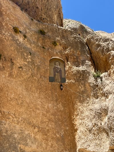 A religious mosaic is affixed to the exterior of a rock wall in the city of Ma'loula, Syria. The mosaic depicts a robed figure with a halo. A small light is suspended above it by a wire. The rock wall is light brown, textured and uneven. The sky is blue in the upper left corner. Small patches of green vegetation are visible on the rock surface.