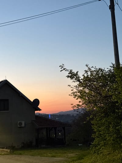 The image captures an evening scene in what appears to be a residential area in Vrnjacka Banja Municipality, Serbia. The sky transitions from a light blue at the top left to a gradient of orange and red towards the horizon line, suggesting a sunset or sunrise. A utility pole and wires are visible in the upper left corner. A leafy tree branch partially obscures the view in the upper right corner. A house stands in the foreground. The house has a dark roof, a satellite dish, a window, and an air conditioning unit. In the background, there is an outdoor seating area with lights. In the far background, there is a hint of a mountainous landscape with a blue light structure. A road and grass are on the right.