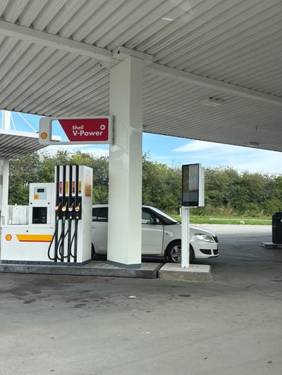 The image shows a Shell V-Power gas station in Sorø, Denmark. A white car is parked at a fuel pump, with the driver's side door open. A digital advertising screen displays the word "salt?". The gas station canopy is visible overhead, along with the blue sky and surrounding trees.