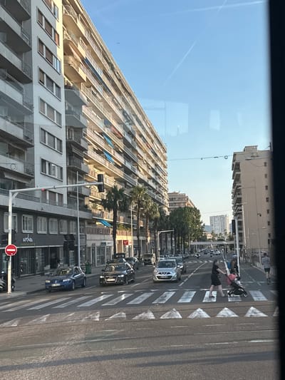 The image depicts a street scene in Toulon, France, likely captured from a vehicle, showing a wide street with multiple lanes of traffic and a pedestrian crossing. On the left, a tall, modern apartment building with balconies dominates the frame, casting a shadow over the street. Across the street, another building of similar height stands. Several cars are visible on the road, including a blue Renault on the left side of the pedestrian crossing and a black Audi to its right. A woman is pushing a stroller across the pedestrian crossing, accompanied by another person. Palm trees line the sidewalks, characteristic of a Mediterranean climate. The sky is clear and blue, with faint contrails from an aircraft visible. The overall impression is of a sunny day in an urban coastal city.