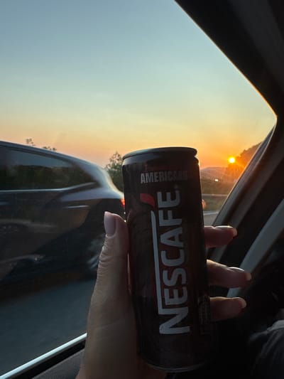 A person's hand, with neatly manicured nails, holds a dark-colored can of Nescafé Americano. The can features prominent white text spelling out "NESCAFÉ" and "AMERICANO". The background is a view through a car window during what appears to be sunset. The sky is a gradient of pale blue to warm orange, with the sun visible as a bright orb. Trees and distant scenery are visible outside the window, suggesting a rural or suburban setting. The overall mood is calm and perhaps contemplative, enhanced by the warm, natural light of the setting sun.