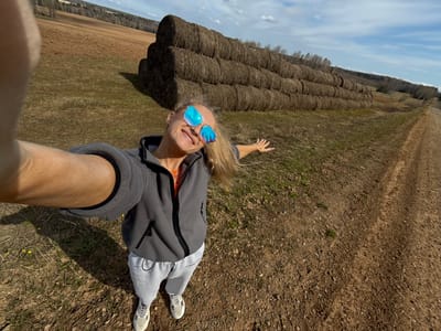 A high-angle selfie captures a smiling blonde woman lying on dry, gravelly ground, looking upwards towards the camera held by her outstretched right arm. She is wearing blue reflective sunglasses, a dark grey zip-up jacket over a lighter top, light grey sweatpants with "STRAND UP" faintly visible on the left leg, and light-colored sneakers. Her body is oriented horizontally across the frame, with her left arm casually extended to the side, suggesting a relaxed pose in an open field.
The ground she lies on is a mixture of earth, small stones, and sparse patches of dry grass. Behind her, a long, neatly stacked row of dark brown round hay bales stretches horizontally across the midground of the scene, indicating an agricultural setting. Beyond the hay bales, a flat, light brown field extends to the horizon, where a dense line of trees forms a dark silhouette against the sky. The upper portion of the image reveals a clear, bright blue sky with scattered white clouds, characteristic of a sunny day, likely midday or afternoon.
The image conveys a sense of outdoor leisure and joy in a rural landscape, consistent with the location context of Малая Уса, Russia.