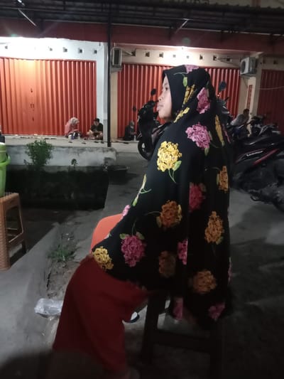 The image shows a side profile of a young woman wearing a black hijab with a floral pattern featuring pink, yellow, and orange flowers. She is seated and looking to the right. She is wearing a red skirt. In the background, there are several motorcycles parked, suggesting an outdoor setting, possibly a street or parking area. Behind the motorcycles, there is a building with large, corrugated red doors, typical of shops or businesses in Indonesia.
Further in the background, other people are visible, some sitting on the ground or a low ledge in front of the building. One person is wearing a light-colored hijab and appears to be interacting with another person seated next to them. Another person is seated in the middle ground, wearing a dark shirt. The lighting suggests it is nighttime or dusk, with artificial lights illuminating the scene. A green bucket is visible in the foreground on the left, and a brown woven stool is to its left. A plastic bag lies on the ground in the lower left portion of the image. The overall scene appears to be a casual gathering or waiting area outside a commercial establishment in Sekopek Wetan, Indonesia. There is no visible text in the image.
