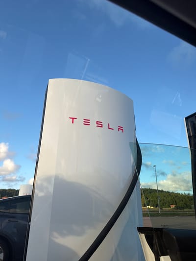 A Tesla Supercharger station is visible in the foreground under a blue sky with scattered clouds. The white, curved structure of the charging station features the prominent red Tesla logo. To the left, a dark-colored Tesla electric vehicle is parked. In the background, a tree-covered hillside is visible with a few buildings. The scene appears to be a public charging area. The image is taken from inside a vehicle, as suggested by reflections on a glass surface. The location is identified as Hogstorp, Sweden.