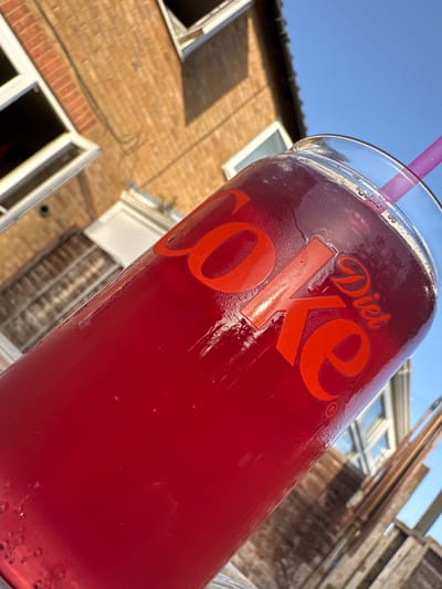 The image shows a glass of dark red Diet Coke with a purple straw, likely taken outdoors in Desborough, United Kingdom, based on the presence of typical British brick houses with white-framed windows. The glass is slightly angled, displaying condensation on its surface. The Diet Coke logo is visible on the glass in red lettering. The background features a brick house and wooden fencing, suggesting a residential garden or patio setting. The sky is blue and clear, indicating sunny weather.