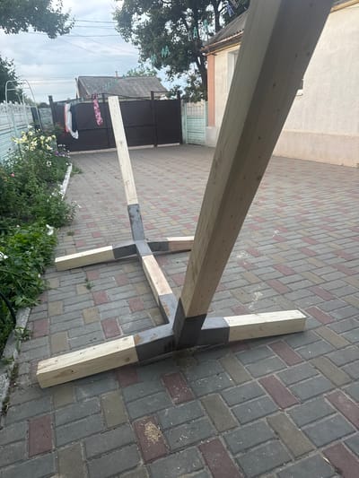 In Heniivka, Ukraine, an unfinished wooden structure, likely a base for an umbrella or some other outdoor fixture, is positioned on a paved driveway. The structure consists of several wooden beams connected by a central metal cross-brace. Two larger beams are angled upwards, supporting a smaller beam that rests horizontally. Four additional wooden beams extend outwards from the metal base, forming a cross shape at ground level. To the left of the structure, a garden bed with flowering plants and greenery is visible. In the background, a dark metal gate and a light-colored house with a tiled roof are situated. Laundry hangs on a clothesline strung between poles in front of the house. The sky above is overcast.
