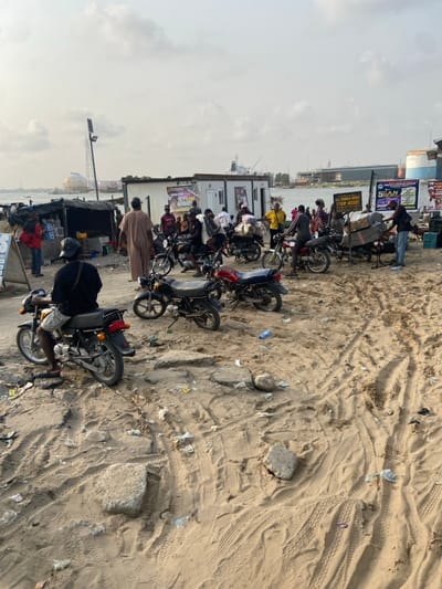 The scene displays a row of motorcycles parked in the sand near a body of water. People stand and gather around the bikes, while a man is seated on one, presumably about to ride off. Tire tracks crisscross the sandy terrain. Buildings and commercial signs are visible in the background. The sky appears overcast.
