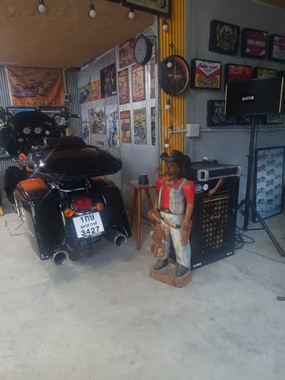 This image was taken indoors in the city of Surat Thani City Municipality, Thailand. The photograph features a black motorcycle in the foreground. To the right of the motorcycle is a cowboy statue dressed in overalls, a red shirt, and a hat. Behind the cowboy statue is a black amplifier and a speaker on a stand. The walls in the background are decorated with various motorcycle-themed posters and memorabilia. The environment appears to be a garage or showroom.