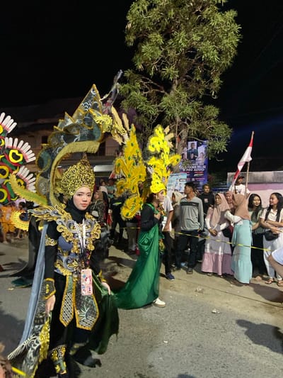This image depicts a nighttime outdoor cultural event or parade on an asphalt street in the city of Kedungbanteng, Indonesia.

In the foreground and mid-ground, two individuals are prominently featured in elaborate costumes. The central figure, positioned left of the frame, is a person with light skin and dark eyes, looking directly at the viewer. This individual wears an intricate, multi-component costume predominantly in black, gold, and dark green. Key elements include a multi-tiered golden crown-like headpiece, ornate shoulder and chest armor with detailed scrollwork, a long dark cape with golden and patterned lining, and a large, arched back structure resembling a stylized dragon or mythical creature, featuring dark green, black, and gold patterns. A smartphone with a light-colored case hangs from a lanyard around their neck.

To the right of the central figure, a second costumed individual wears a prominent yellow, fan-shaped or feather-like headpiece and a long, flowing green garment over a black long-sleeved top. This person is partially obscured and oriented towards the left.

The right side of the street is lined by approximately fifteen spectators, a diverse group of men and women of varying ages, some wearing hijabs, dressed in casual attire. They are positioned behind a yellow rope or tape acting as a crowd barrier, observing the event. One spectator on the far right holds an Indonesian flag on a pole.

The background features a large, leafy tree centrally located. Adjacent to the tree on the right, a large banner is visible, displaying images of individuals and text, including "ANNIVERSARY 2023" and "PRO AUDIO". Dimly lit buildings are present in the far left background. The asphalt street surface shows some scattered light debris. The entire scene is illuminated by artificial light sources, indicating nighttime conditions.