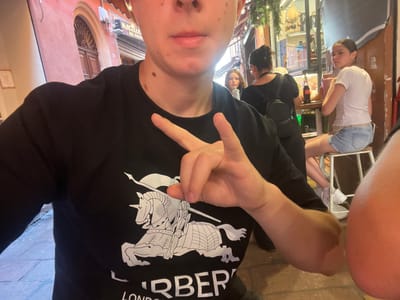 The image appears to be a selfie taken outdoors in Bologna, Italy. The photographer is wearing a black t-shirt with a white graphic design featuring a knight on horseback and the word "London" above it. The photographer is holding up their hand, making a gesture. Behind the photographer, there are other individuals including a girl in a white top and denim shorts seated at a table, and a person wearing a face mask and dark clothing. Shops and buildings with signs, awnings, and decorations are visible in the background. The architectural details and signage suggest a narrow street or alleyway in the old town area of Bologna. A sign is visible on the right that says "Macelleria del Vicolo", which is a business name and could also refer to the name of the street.