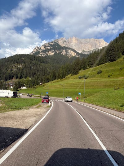 The image captures a scenic mountain road in the vicinity of Mazzin, Italy. A paved road with white lane markings curves through a landscape dominated by lush green hills and a dense pine forest that climbs the mountainside. In the distance, a majestic, rocky mountain peak with sheer cliffs stands against a backdrop of a partly cloudy blue sky.
In the foreground, the road is traveled by a white car in the right lane and a red car parked on the left side of the road, near a gravel shoulder. Further down the road, a pedestrian crossing sign is visible. To the left of the road, a parking area can be seen with several motorcycles and a white trailer. Two individuals, one wearing a red shirt and the other in a yellow vest, are standing near the white car. The environment suggests a summer day with pleasant weather, ideal for outdoor activities in the Dolomites region.