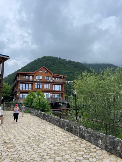 In the city of Abaata, Abkhazia, a hotel with a wooden facade and multiple stories is situated against a lush green mountainside partially obscured by clouds. Two women are walking on a paved pathway in front of the hotel. The pathway is made of interlocking hexagonal and square tiles in shades of yellow and grey. A stone wall borders the pathway on the right, with a metal railing on top. Trees and foliage surround the hotel and the pathway, with some trees bearing string lights. A bridge is visible behind the hotel, spanning a body of water. The sky is overcast with heavy clouds.