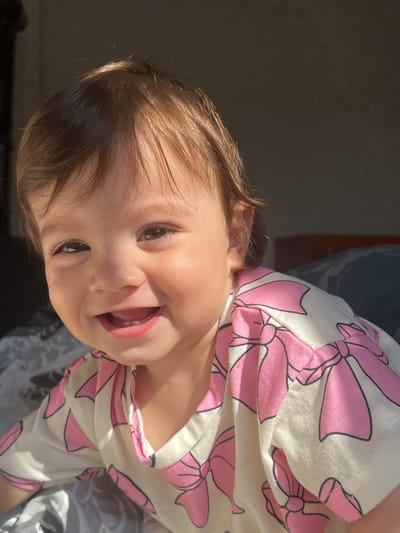 This is a medium close-up, outdoor, eye-level shot of a smiling baby. The baby is facing the viewer, with their head tilted slightly to the left. The baby has light brown hair and is wearing a white t-shirt with pink bows. The background is blurred and appears to be a dimly lit room with sunlight streaming in from the left. There are no specific identifiable landmarks or indications of the city of Elena, Bulgaria in this image.