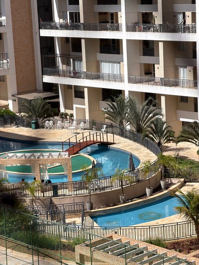 Here's a description of the image:
The image shows an overhead view of a residential area in Brasília, Brazil. There is a tall apartment building to the left of the frame, constructed with light beige walls and white accents. The building features multiple balconies with dark railings.
Adjacent to the building is a swimming pool area. The pool is in a kidney bean shape with a central island. The pool is surrounded by lounge chairs and palm trees. A chain-link fence borders the pool area.