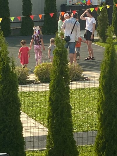 In Novi Petrivtsi, Ukraine, a group of people are gathered outdoors, possibly for an event or celebration, indicated by the colorful triangular flags strung up. The scene is bathed in the warm light of what appears to be late afternoon or early evening. Several individuals are visible, including a person in a white t-shirt and dark shorts, another in a blue and white shirt with a black crossbody bag, and a person in a red dress. They are standing on a paved area adjacent to a well-maintained green space, possibly a lawn or artificial turf, enclosed by a wire mesh fence. Cypress trees or similar greenery line the perimeter of the scene, partially obscuring the view. The overall atmosphere seems casual and relaxed, with people interacting and observing their surroundings. The ground is paved with rectangular tiles. The specific event is unclear, but the presence of decorations and the gathering of people suggest a social occasion.