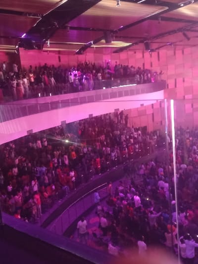 The image captures a vibrant, high-angle view of a large indoor public space, bustling with a significant crowd of people. The perspective is from an elevated position, looking down into a multi-level or expansive area.

On the left side of the frame, there is a prominent vertical wall or architectural facade. It features a pattern of rectangular panels, possibly made of wood or a similar textured material, illuminated with a warm, pinkish-purple light. Darker, vertical beam-like structures are also visible on this wall, with some horizontal elements attached, suggesting structural supports or decorative features.

In the center of the image, a wide, light-colored pathway or ramp descends diagonally from the upper left towards the lower right. This surface appears smooth and reflective, catching the bright pink and white light sources, creating luminous streaks. Along this central pathway, and extending into the broader floor area below, a large number of people are gathered. They appear as small, indistinct figures due to the distance and lighting, but their sheer density indicates a crowded environment. Many figures show splashes of color in their clothing, including reds, whites, and various dark hues.

To the right of the central ramp, a dark, curved structure, possibly a balcony railing or another walkway, parallels the ramp's descent. More people are visible on this side, further contributing to the impression of a packed venue. The overall lighting scheme is dominated by strong pink and purple hues, casting a theatrical or event-like ambiance over the entire scene. Bright white light sources are also present, particularly reflecting off the central ramp and illuminating some areas of the crowd.

The activity appears to be a large gathering or event, with people moving, congregating, or observing something within this large indoor space. The intense lighting and large crowd suggest an evening or night-time setting for a public spectacle, concert, or a very busy retail/entertainment complex. No specific text is visible in the image.