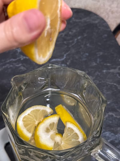 A hand is shown squeezing a lemon slice over a glass pitcher filled with water and lemon slices. The pitcher has a faceted design and is made of clear glass. The surface the pitcher is on appears to be a dark, textured countertop. The background is blurred, but suggests an indoor setting. The scene is focused on the action of preparing a lemon-infused drink. There is no information about the location in Raivka, Ukraine.