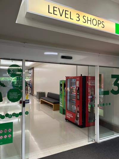 This image captures an interior view of a well-lit commercial space, likely a shopping center or public building corridor, identified as "Level 3 Shops" in Indooroopilly, Australia. The scene is presented from a slightly elevated and angled perspective, looking into a lounge or waiting area through a glass partition.

On the left, a bright, rectangular sign with internal illumination prominently displays "LEVEL 3 SHOPS" in capital letters, indicating the floor and general purpose of the area. Below this sign, a small black security device is mounted on a light-colored frame.

The central and right portions of the image feature a glass wall separating the foreground from a seating area. Within this area, two vending machines are visible: a large red Coca-Cola machine adorned with "REFRESH HERE" branding and images of drinks, and a smaller green machine clearly labeled "Pay here." In front of these machines, a contemporary grey couch with a light wooden base offers seating. The flooring is a polished, light-colored surface, possibly tile or polished concrete, transitioning to a darker carpeted area further into the background.

The glass partition is decorated with several green circular decals containing information and symbols. Large text includes "3 hours FREE parking," "FREE when you enter after 5PM every day," and "No ticket required, just remember your rego," detailing parking instructions. A prominent green "L3" is also visible on the glass, reiterating the level. Other smaller green icons, likely conveying rules (e.g., no dogs, no littering), are scattered across the glass.

The overall atmosphere is quiet and functional, with no individuals visible in the immediate area. The setting appears modern, clean, and designed for visitor convenience, offering refreshments and a place to rest while providing clear directional and parking information.