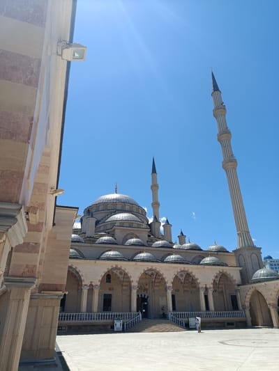 A wide-angle, low-angle shot captures the exterior of the Akhmad Kadyrov Mosque in Grozny, Russia, under a clear blue sky with a bright sun casting light rays. The mosque features a large central dome and multiple smaller domes, all with ribbed metallic surfaces. Two tall, slender minarets with ribbed shafts and decorative balconies rise from the mosque complex. The mosque is constructed with light-colored stone, accented with darker pinkish-brown banding on the pillars and around some of the arched windows and doorways.

In the foreground, to the left, is a section of an adjacent building with a stone facade displaying distinct pinkish-brown stripes and vertical paneling. A white exterior light fixture is mounted on the upper portion of this building. A courtyard area, paved with light-colored stone, stretches in front of the mosque. A white balustrade with vertical railings runs along a raised section in front of the mosque's arched facade, which features multiple arcades supported by columns.

Two individuals, dressed in light-colored clothing, are standing near the entrance of the mosque on the courtyard level, appearing to be interacting or conversing. One person is wearing a white shirt and light-colored pants, while the other is wearing a white shirt and darker pants. A sign with figures and text is visible near the stairs leading up to the mosque. In the background, a portion of a modern building with a blue dome and windows can be seen. The overall scene depicts a sunny day at a prominent religious site.