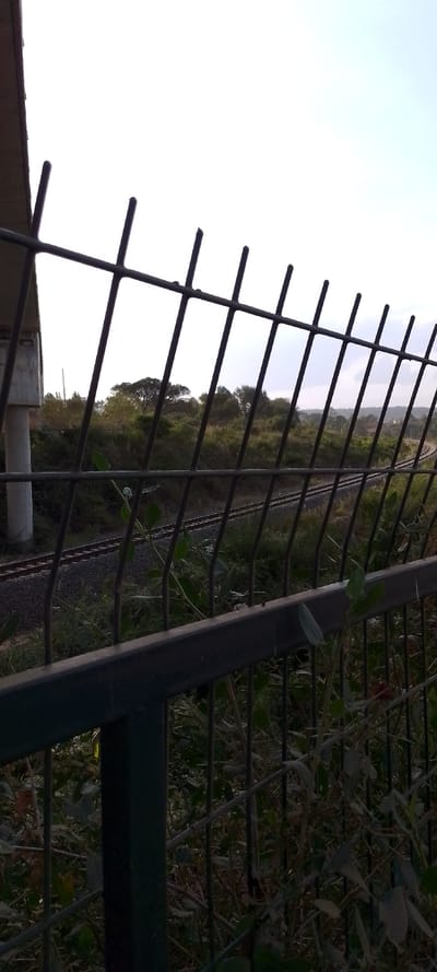 A metal fence dominates the foreground, partially obscuring a railway track winding through overgrown vegetation. A concrete bridge pillar is visible to the left. Trees and shrubs line the track, while the sky is bright and overcast. The overall scene suggests a rural or semi-rural setting with infrastructure elements blending into the natural environment.