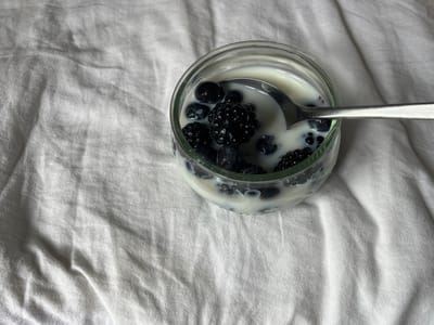 A close-up, high-angle shot features a clear glass container filled with a white liquid, likely milk or yogurt, mixed with fresh dark berries. Inside the glass, numerous dark blueberries and several textured blackberries are visible, some partially submerged in the liquid. A silver metal spoon rests within the container, its bowl immersed in the liquid and berries, suggesting it is either being prepared or consumed. The background consists entirely of a soft, rumpled white fabric, possibly a bedsheet or tablecloth, which creates a textured and minimalist setting for the food item. The folds and shadows in the fabric add depth to the otherwise simple backdrop. The scene evokes a tranquil moment, possibly a breakfast or a healthy snack. No individuals are present in the image. The lighting appears natural, suggesting daytime. There are no visible location cues within the image itself beyond the given context of Aarhus, Denmark, and no discernible text.