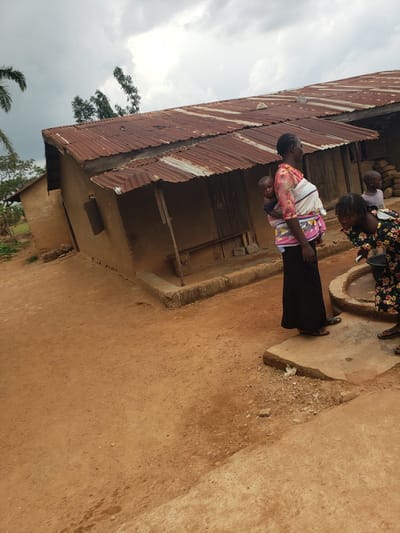 In Kachia, Nigeria, a scene unfolds with a focus on daily life and community interaction. A rustic, mud-walled structure with a corrugated iron roof dominates the background, its weathered appearance suggesting a long-standing presence in the environment. The roof, showing signs of age and repair, is a testament to the building's history.
In the foreground, several individuals are engaged in various activities. A woman, her hair neatly braided, carries a baby on her back, a common practice in many cultures. She is dressed in a vibrant, patterned top and a long, dark skirt. Beside her, another young woman in a colorful, floral dress is bent over, seemingly interacting with something out of frame or on the ground. Another child stands nearby, looking towards the right side of the image.
The ground is a dusty, reddish-brown earth, typical of the region, with some scattered stones and a small, concrete-like platform. The atmosphere appears overcast, with a cloudy sky suggesting potential for rain. The overall impression is one of a simple, rural setting, where community and family are central to the depicted activities. The image captures a moment of everyday life in Kachia, providing a glimpse into the lives of its residents.