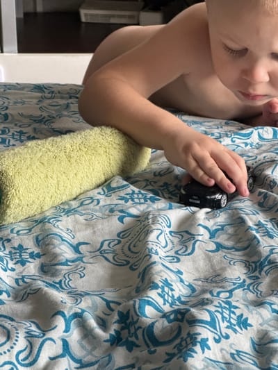 A close-up shot captures a young child's head and arm as they interact with a miniature black car. The child, appearing to be a toddler, is seen from the side, with their face partially obscured and their eyes closed or looking down. Their delicate eyelashes are visible. The child's bare shoulder is in frame, and their arm is extended, holding the toy car. The car is black with silver accents on the wheels and what appears to be a logo or emblem on its side.
The child is nestled against a textured surface that resembles a soft, pale yellow towel or blanket. Behind the child and the towel is fabric with a repeating pattern of blue, stylized floral or paisley designs on a white background. The fabric is draped and creased, suggesting it might be a bedspread or upholstery.
The overall mood is intimate and tranquil, possibly indicating a moment of play or rest. The lighting appears to be indoor, with no strong indicators of the time of day or specific weather conditions. The background is blurred, but hints of what might be furniture or appliances in a domestic setting are discernible on the left side of the image. There is no visible text in the image. The scene evokes a sense of childhood innocence and gentle interaction with toys.