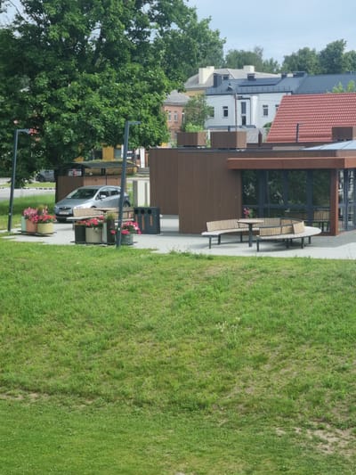 This is a photograph of an outdoor public area in Kuršėnai, Lithuania, likely near a park or community space. In the foreground, a gently sloping grassy area dominates the lower half of the frame. The middle ground features a paved area with several objects. A silver sedan is parked in front of a covered structure with wooden slatted walls. To the left of the car are planters filled with pink and white flowers. To the right of the car, there are three recycling bins. Further to the right, a modern building with large windows and brown wooden siding is visible, with a curved wooden bench and table in front of it. In the background, trees and buildings are present, suggesting a town setting. A large tree with dense green foliage is prominent on the left side of the image. The sky is overcast. There are no people visible in the photograph.