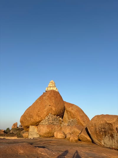 The image shows a view of the Hanuman Temple at Hampi, located in Kamalapura, India. The temple is built on top of a large rock formation. The sky is a clear blue with a slight gradient. The rock formations are primarily reddish-brown. In some parts, one can see evidence of structures made from stones that might be linked to the temple. The sun is on the rocks, creating shadows on the rock surfaces.