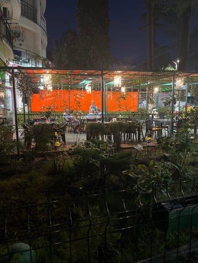 The image depicts an outdoor seating area of a restaurant or cafe at night. The scene is illuminated by warm, ambient lighting from decorative string lights and hanging lamps, casting a soft glow on the foliage and tables. Several people are seated at tables, engaged in conversation and enjoying the evening. The setting appears to be lush with plants and greenery, creating a cozy and inviting atmosphere. In the background, there are glimpses of buildings and a dark night sky. A metal fence is visible in the foreground, suggesting the edge of the property. The overall mood is relaxed and convivial. There are no clearly discernible signs or text visible in the image.