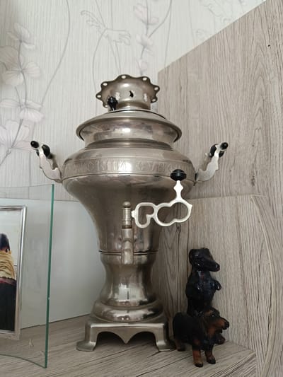 A silver samovar sits on a shelf next to a picture frame and two dachshund figurines. The samovar has inscription in Russian which reads: “ТУЛЬСКИЙ ОВЧА". The wallpaper behind the samovar is light-colored with floral pattern. The shelf appears to be made of a light wood. The photograph is likely from an indoor location. The exact location in Serpukhov, Russia cannot be definitively determined from the image alone.