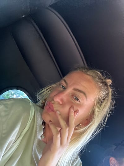 A blonde woman poses inside a dark vehicle. She's angled towards the camera with a slight pout. Her hair is partially up, and she wears a light-colored shirt, gold hoop earrings, and a necklace. Her hand is near her face, displaying short, manicured nails. The interior is dark, possibly leather or vinyl, with a small, round window visible in the background showing a glimpse of greenery. The lighting is bright, suggesting daytime.
