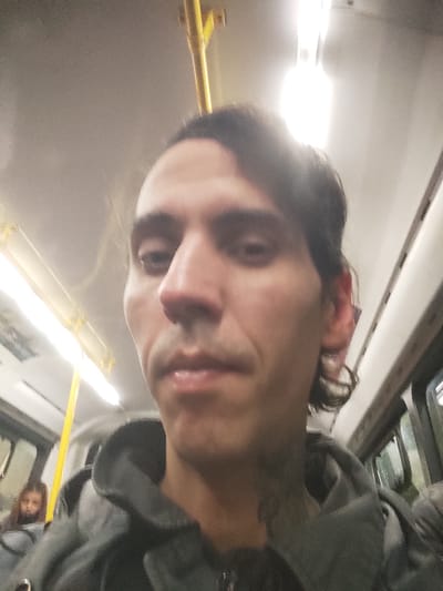 A man with dark hair and a gray hoodie is taking a selfie on a bus. Another person is visible behind him on the left. The bus interior is illuminated by overhead fluorescent lights. Based on the location given in the prompt, the bus is assumed to be in Gerli, Argentina.