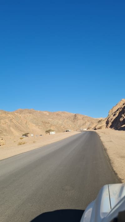 The image depicts a desert road scene near Eilat, Israel. The road, made of dark asphalt, curves along a mountainside with arid, rocky terrain. The sky is clear and bright blue. Several small buildings, likely residences or rest stops, are visible near the base of the mountain alongside the road. The side of a light-colored vehicle is in the right of the image, implying the photo was taken from inside the car.