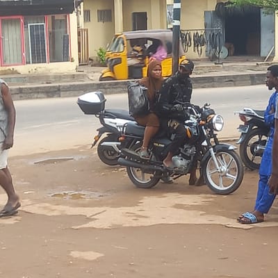 A woman wearing a hijab is riding on the back of a motorcycle in the city of Sardauna, Nigeria. The motorcycle is parked on a street next to a three-wheeled vehicle. Two men are standing nearby. The buildings on the street appear to be shops or residences.