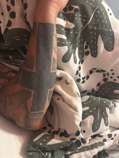 This close-up shot features a person's arm, heavily adorned with a blackwork tattoo, resting amidst a patterned blanket. The arm, exhibiting a medium skin tone, is primarily covered by a bold, geometric tattoo that creates large, solid black shapes against the skin, possibly forming a cross-like design or an abstract pattern with defined edges.
The blanket, likely a duvet or comforter, has a light cream or white background and a distinct, repeating pattern. This pattern includes large, dark green, abstract organic shapes that resemble stylized leaves or cacti, detailed with thin white lines and scattered black dots. Another element of the blanket's design involves lighter beige or tan shapes with prominent black spots, giving the impression of a spotted animal print. The fabric of the blanket appears soft and has visible folds and creases, indicating it is draped around the arm or body.
The setting appears to be indoors, possibly a bedroom, suggesting a moment of rest or relaxation. The lighting is soft and even, creating a cozy atmosphere without harsh shadows. No specific time of day is discernible from the light. There is no visible text in the image. The location context provided is Vladaya, Bulgaria.