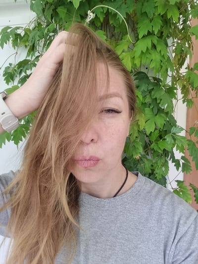 A woman is lying on her side with her head resting on her hand. She has light brown hair that is partially covering her face. She is wearing a gray t-shirt. Behind her is a wall covered in green leafy vines. The location is not identifiable.