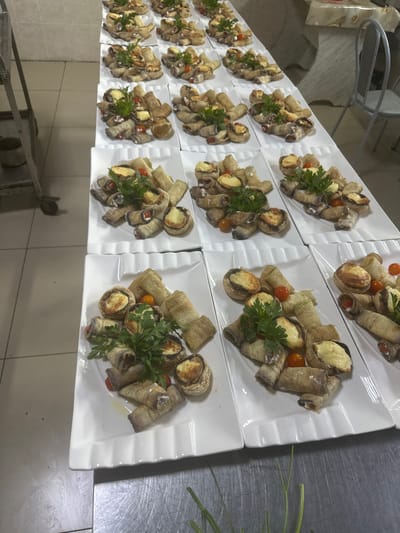 The image shows a catering setup with multiple white rectangular plates filled with appetizers. Each plate contains several pieces of what appear to be stuffed eggplant rolls, some stuffed mushrooms with a golden-brown topping, and small cherry tomatoes. Fresh parsley sprigs are used as a garnish on many of the plates. The plates are arranged in rows on a white surface, likely a table or counter, and some are partially obscured by others. In the background, a stainless steel kitchen cart is visible to the left, and chairs are present to the right. The overall impression is that of a prepared meal for an event. There are no people or specific landmarks visible that would indicate the city of Kostanay, Kazakhstan, or any other specific location. The environment appears to be an indoor kitchen or preparation area.