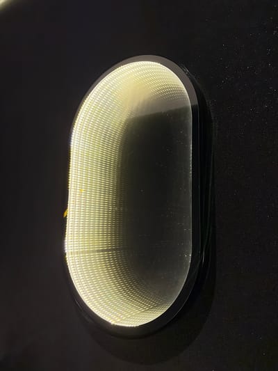 An oval-shaped, illuminated object, possibly a mirror with an infinity effect, is mounted on a dark, textured wall. The object emits a bright, warm yellow light from within, creating a tunnel-like visual effect with repeating patterns of light and reflection. The wall appears to be painted black or a very dark gray, with subtle speckling or texture. The lighting conditions suggest it is either nighttime or an indoor setting with low ambient light, highlighting the illuminated object. No people or actions are visible. The object is centrally located in the frame, with a portion of the dark wall visible on either side. The setting is unknown, but the description specifies the location as Baan Lipa Noi, Thailand.