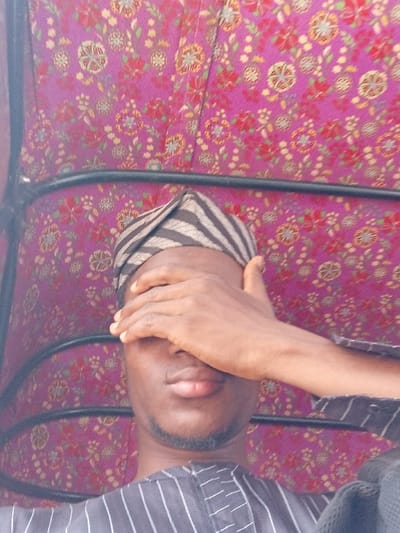 In Katcha, Nigeria, a medium close-up shot depicts a male individual within an enclosed space. The subject has dark skin, short facial hair on his chin and jawline, and wears a horizontally striped cap (black and light-colored) along with a vertically striped dark-colored garment. His right hand is raised, palm inward, covering his eyes and upper nasal area. The background features a magenta-colored fabric lining patterned with repeating floral motifs in red, green, gold, and white. A black, curved metal bar, indicative of a structural support, is visible in the upper portion of the frame, suggesting the interior of a vehicle, possibly a motorized tricycle.