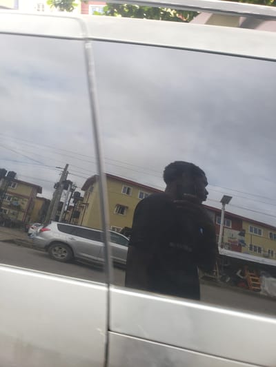 A reflection in the window of a silver vehicle shows a man in a black t-shirt holding a dark mobile phone to his right ear, suggesting he is on a call. The man's head is tilted slightly down and to his right, and his eyes are not visible. Behind him, the reflection shows a street scene in Iganmu, Nigeria, with a yellow building with red trim, several utility poles with wires, and a silver SUV parked on the side of the road. The sky is overcast, and trees with green leaves are visible at the top of the reflection. The image is taken from the perspective of someone standing next to the silver vehicle, looking at its reflective window.