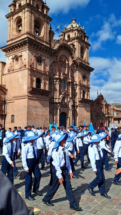 This image captures a procession in the Plaza de Armas in Cuzco, Peru, in front of the Cusco Cathedral. A group of young people, primarily boys, are marching in uniform. They are wearing white long-sleeved shirts, dark ties, dark pants, and matching blue hats with wide brims. Some are also wearing dark jackets or vests over their shirts. They appear to be part of an academic or cadet program, judging by their organized formation and uniforms.
Behind the marching group, the magnificent facade of the Cusco Cathedral dominates the background. The cathedral, a historic colonial-era church, is constructed of reddish-brown stone and features intricate architectural details, including arched doorways, ornate carvings, and towering bell towers. A rainbow flag is visible flying from one of the cathedral's spires, adding a touch of color to the scene. The sky above is a bright blue with scattered white clouds, suggesting a clear and sunny day. The ground is paved with cobblestones, typical of historic city squares. Other individuals, presumably spectators or participants, are visible in the background, some holding flags or banners. The overall atmosphere appears to be one of a formal event or parade.