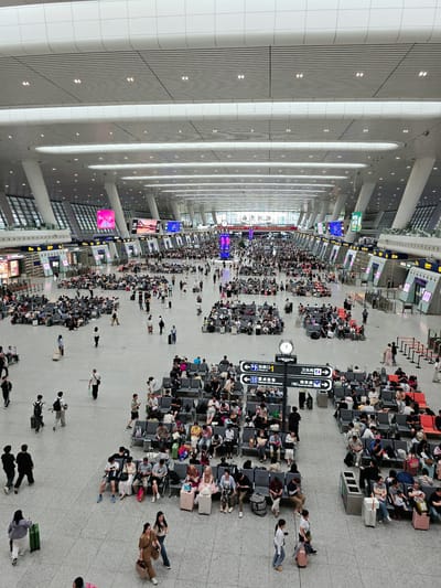This image captures a wide-angle view of the interior of a bustling train station in Shangcheng District, China. The vast, modern space features a high, illuminated ceiling with a patterned design and large supporting pillars. Numerous digital displays are visible throughout, showing information like track numbers and destination boards, some with Chinese characters. The floor is a light-colored, tiled surface.
The main area is filled with a large crowd of people waiting, either standing or seated on rows of grey benches arranged in sections. Many individuals are carrying luggage, including suitcases, backpacks, and tote bags. People are seen walking, talking, looking at their phones, or attending to their belongings. Some are dressed in casual attire, while others appear to be more formally dressed.
Several boarding gates are visible, indicated by signage with numbers and letters. The overall atmosphere suggests a busy travel hub during a peak period. In the foreground, there are directional signs for "Check-in," "Passengers with Special Needs," and "Business Class," as well as for "Restrooms." The presence of a Longines clock on a sign adds a detail of timekeeping. The scene is well-lit, with artificial lighting from the ceiling and the digital displays contributing to the brightness.