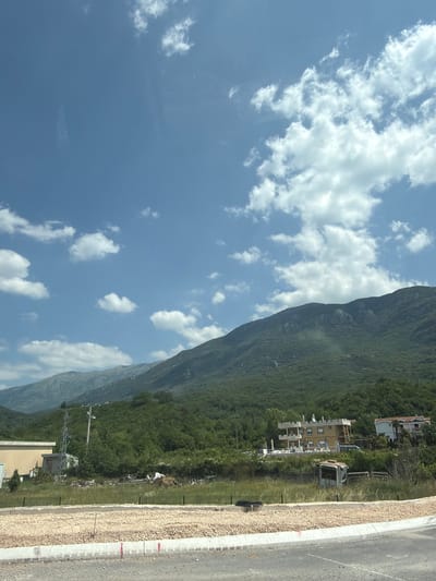 The image depicts a scenic landscape in Kovačko Polje, Montenegro, under a bright blue sky with scattered white clouds. In the foreground, a road with a gravel embankment and a concrete curb is visible. To the left, a light-colored building is partially shown. In the middle ground, a grassy area contains some scattered debris, including what appears to be discarded chairs and a pile of white material. A tall metal tower stands next to a smaller concrete structure. Further back, several buildings are situated on a hillside, including a multi-story building with a light yellow facade and a white tiled roof, and a white building with an orange-tiled roof. The background is dominated by a lush green, forested mountain range. The overall scene suggests a rural or semi-rural environment with some ongoing construction or development.