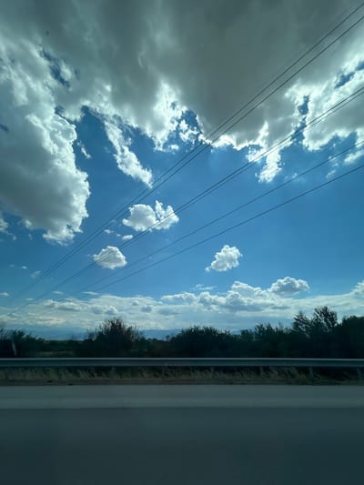 This image depicts a wide-angle, outdoor scene. The sky dominates the upper portion of the frame, filled with a dramatic display of fluffy white and grey clouds against a vibrant blue backdrop. Several power lines crisscross the sky diagonally. Below the sky, a band of dark green trees and vegetation forms a natural horizon. In the foreground, a grey asphalt road is visible, separated from the landscape by a metal guardrail. The scene is captured from a moving vehicle, suggesting a roadside perspective. No people or specific buildings are identifiable, and the location in Shtarkovo, Bulgaria is not visually evident within the image itself. The overall impression is one of a dynamic sky over a natural, undeveloped landscape.