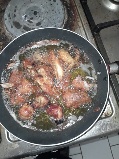 A close-up, overhead shot shows pieces of seasoned meat frying in a black frying pan filled with hot oil. The meat, likely chicken, is golden brown and appears to be seasoned with spices. Bubbles and foam surround the meat as it fries. The pan is on a dirty, stained metal stovetop. The stovetop is situated on a counter, with a tiled floor visible in the lower part of the frame. The overall scene suggests a meal being prepared at a residence in Dam Camp, Nigeria.