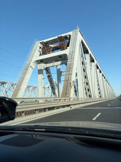 The image captures a detailed view of the Borcea Bridge in Borcea, Romania, from the interior of a vehicle traveling on its highway section. The foreground features the dark dashboard and lower windshield of a vehicle, with a black device or mount visible on the dashboard's left side.
The primary subject is a massive, light-grey steel truss bridge, which accommodates both a multi-lane highway and an electrified railway. The highway pavement, appearing as dark asphalt with a distinct white dashed lane marking and a solid white edge line, occupies the mid-ground. Grey metal guardrails run parallel to the road, separating it from the main bridge structure.
The main bridge structure, a large steel truss, dominates the right side and upper half of the image. It exhibits a complex geometric design with riveted connections and integrated access ladders on its exterior. Within the upper section of the closest truss, internal support elements, appearing darker and possibly corroded, are visible. To the left of the highway, a separate, older-looking grey steel truss railway bridge runs parallel, equipped with overhead electrical catenary lines. The sky above is uniformly clear and bright blue, indicating sunny daytime conditions. No individuals are present in the scene.